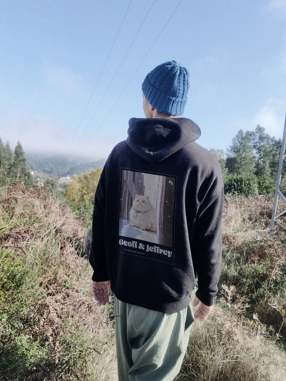 Man wearing black organic cotton Geoff & Jeffrey hooded sweater with drop shoulders  and a large graphic backprint of a joyful, fat cat in the snowy city with the brandname in ohno blazeface and title in libre baskerville printed below.