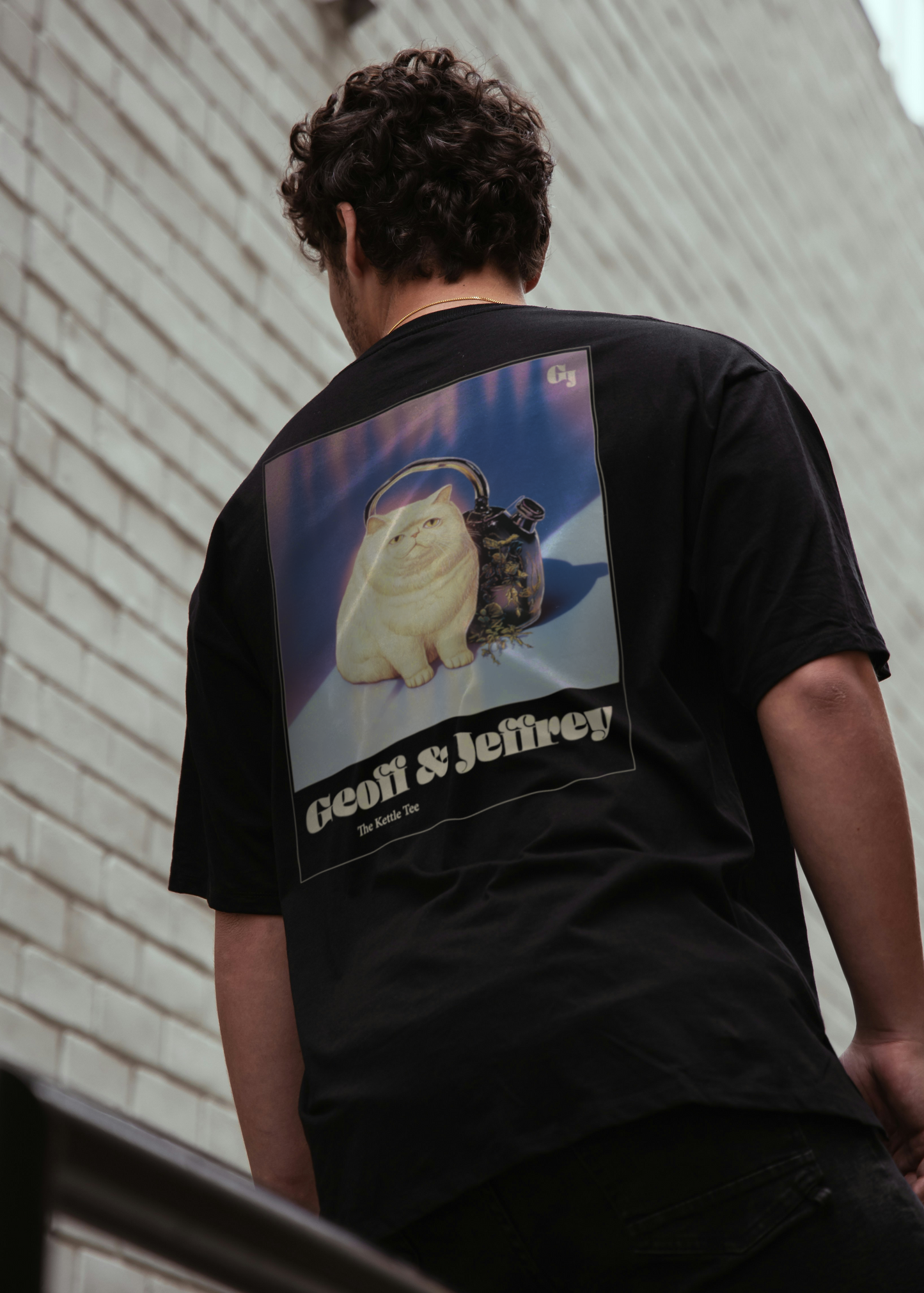 Man wearing a black organic cotton Geoff & Jeffrey unisex t-shirt with drop shoulders  and a large graphic backprint of the weird cat Jeffrey with his kettle. with the brandname and title printed below. The t-shirt has a cool aesthetic.