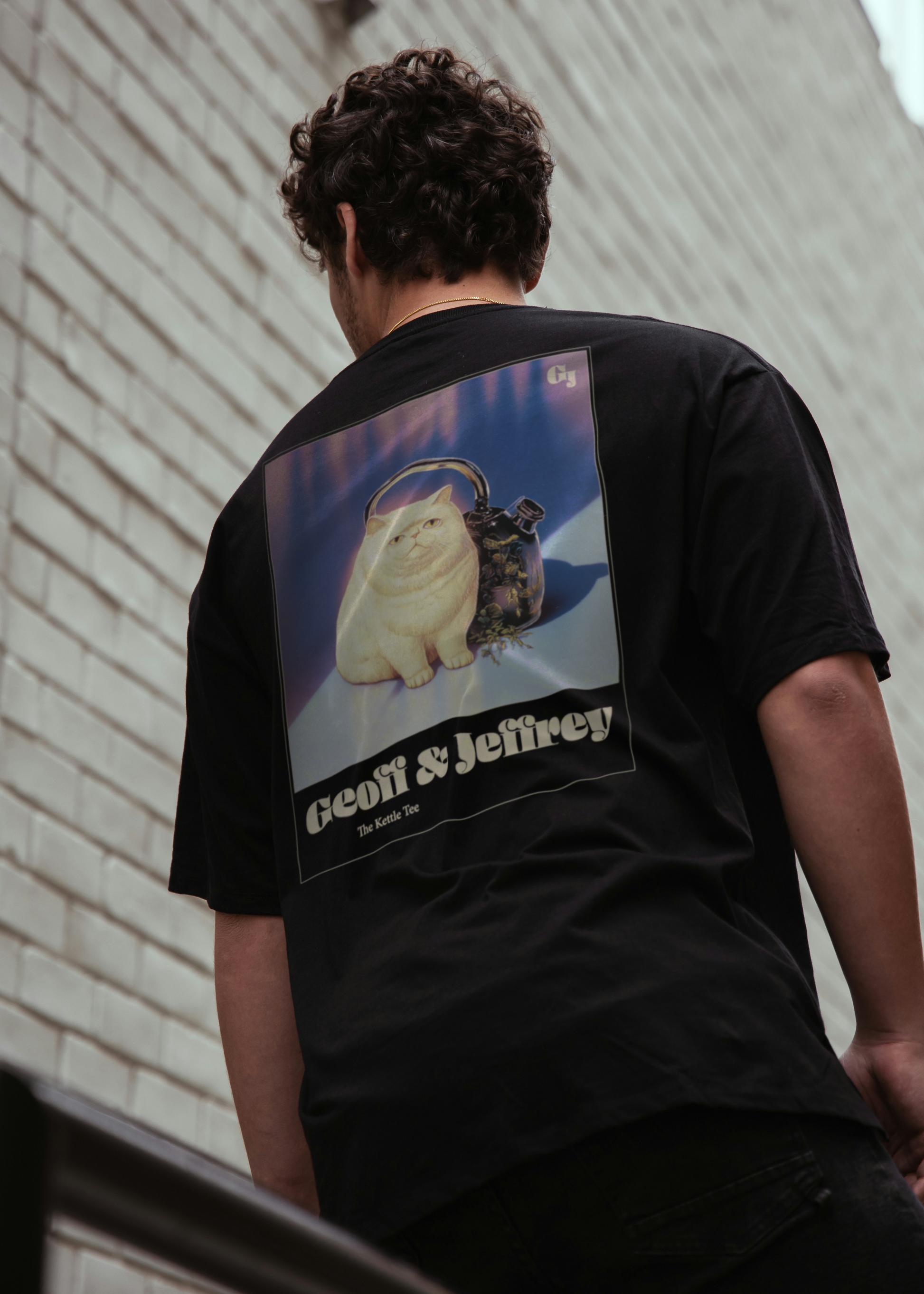 Man wearing a black organic cotton Geoff & Jeffrey unisex t-shirt with drop shoulders  and a large graphic backprint of the weird cat Jeffrey with his kettle. with the brandname and title printed below. The t-shirt has a cool aesthetic.
