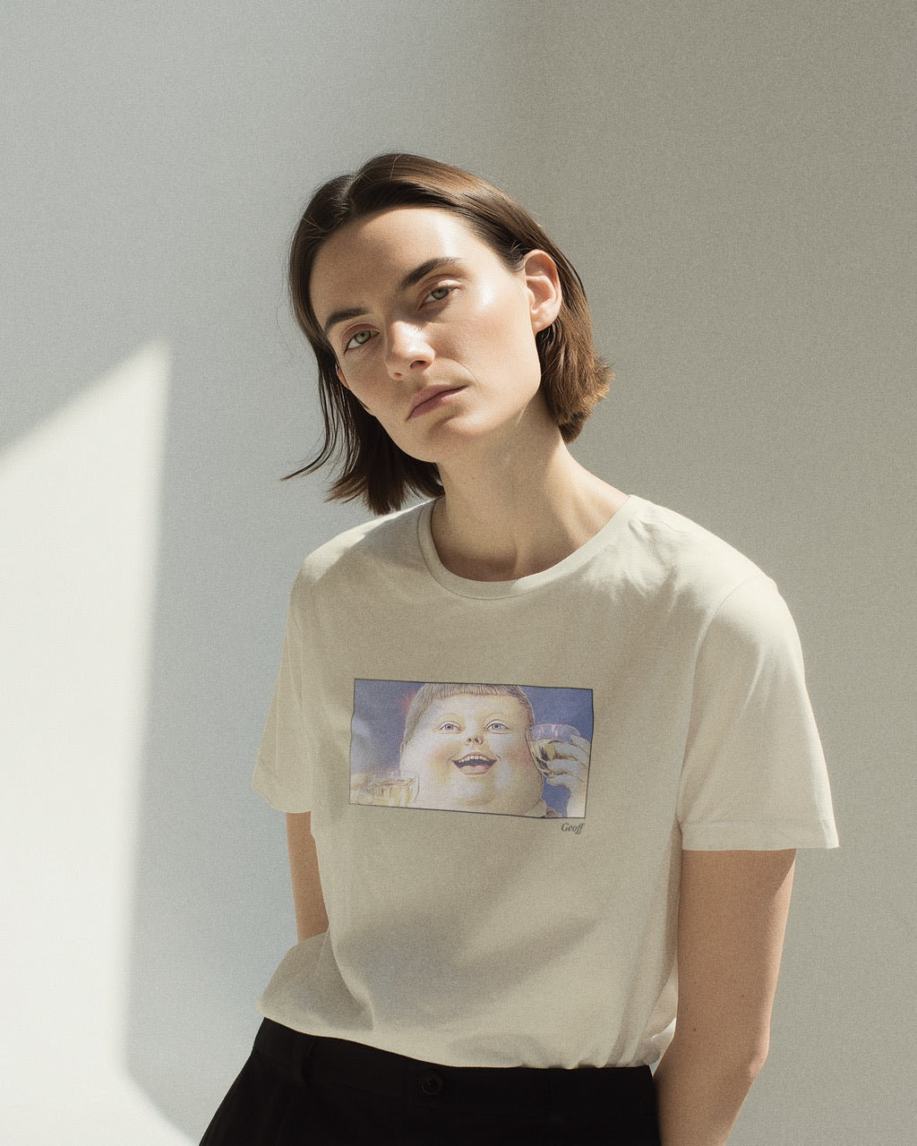 Model in a women's off-white natural organic cotton Geoff & Jeffrey t-shirt with a framed graphic print of a jolly, proud boy (Geoff) looking upwards holding drinks for apero, with his name 'Geoff' subtly printed in Libre Baskerville below the image. The t-shirt has a feel good aesthetic.