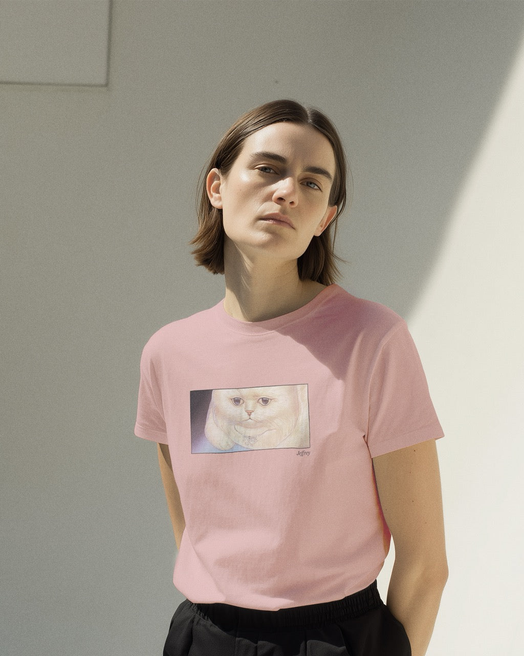 Model in a women's soft pink organic cotton Geoff & Jeffrey t-shirt with a framed graphic print of a funny fat cat (Jeffrey) looking worried. With his name 'Jeffrey' subtly printed in Libre Baskerville below the image. The t-shirt has a cute and dreamy aesthetic.
