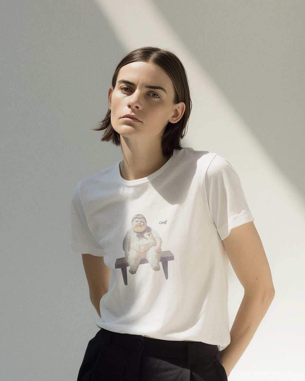 Model wearing a white organic cotton Geoff & Jeffrey t-shirt with a graphic print of a jolly, proud boy (Geoff) sitting on a bench, waiting, with his name 'Geoff' subtly printed in Libre Baskerville next to the image. The t-shirt has a weird aesthetic