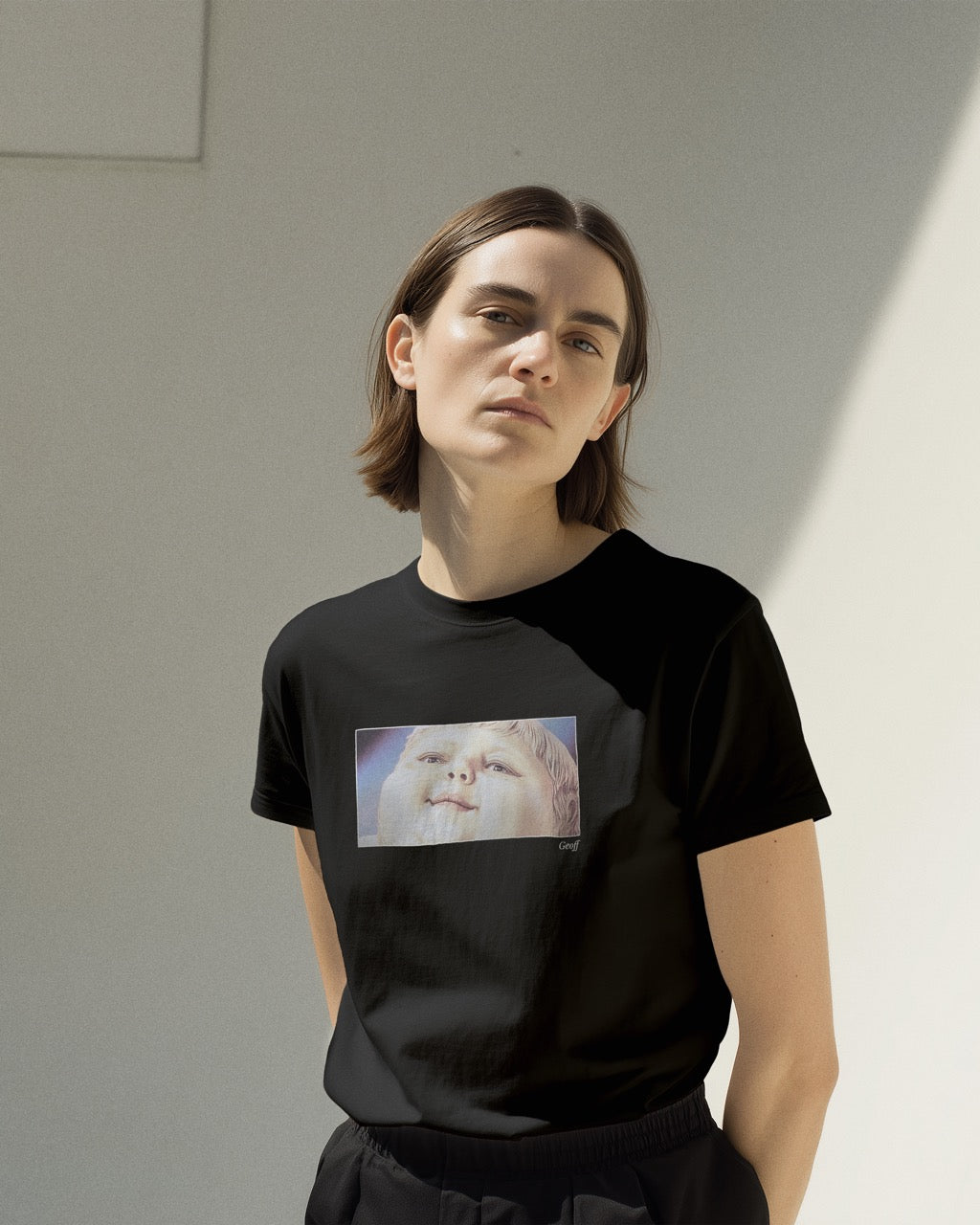 model in women's black organic cotton Geoff & Jeffrey t-shirt with a framed graphic print of a jolly, proud boy (Geoff) looking upwards, smiling, with his name 'Geoff' subtly printed in Libre Baskerville below the image. The t-shirt has a feel good aesthetic