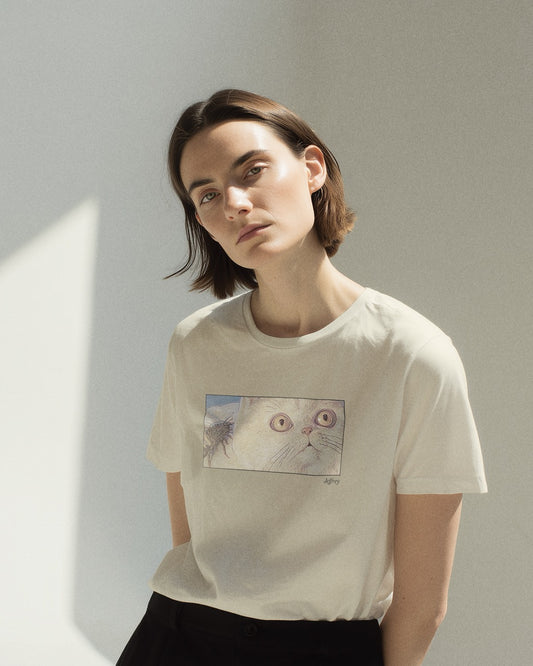 Model in women's off-white natural organic cotton Geoff & Jeffrey t-shirt with a framed graphic print of a funny fat cat (Jeffrey) looking concerned with a wasp on his back. With his name 'Jeffrey' subtly printed in Libre Baskerville below the image. The t-shirt has a cute aesthetic