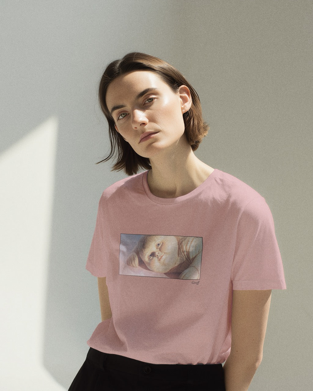 Model in women's soft pink organic cotton Geoff & Jeffrey t-shirt with a framed graphic print of a jolly, proud boy (Geoff) looking slightly upwards, with his name 'Geoff' subtly printed in Libre Baskerville below the image. The t-shirt has a dreamy aesthetic