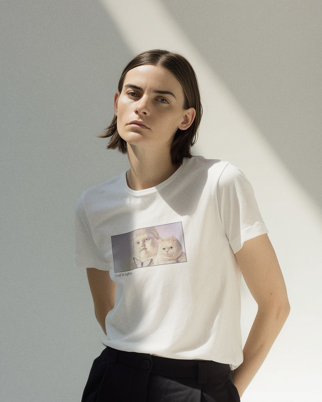 Model in a women's white organic cotton Geoff & Jeffrey t-shirt with a framed graphic print of the jolly, proud boy (Geoff) and his funny fat cat (Jeffrey), holding a butterfly, with there names subtly printed in Libre Baskerville below the image. The t-shirt has a dreamy aesthetic.